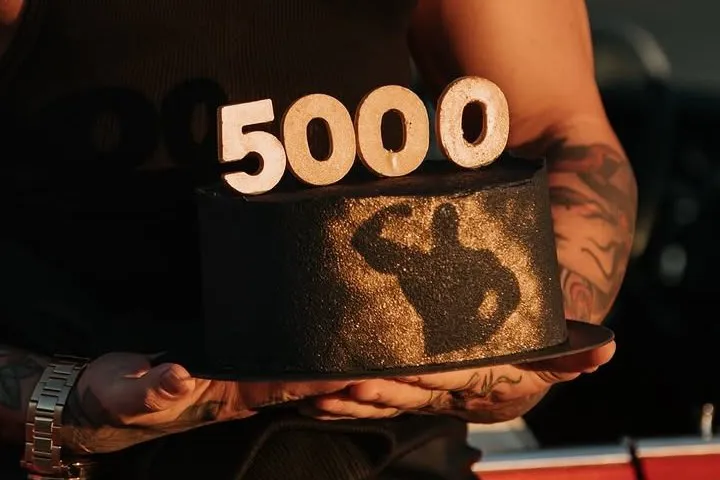 Melna kūka ar zelta siluetu un baltiem cipariem „5000” saulrieta gaismā